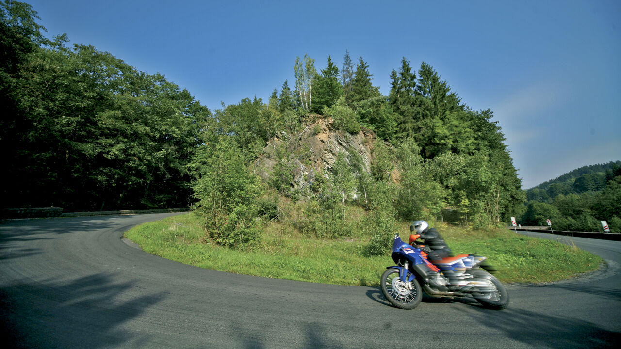 Reiseenduro in einer weiten Rechtskurve vor bewaldetem Hang